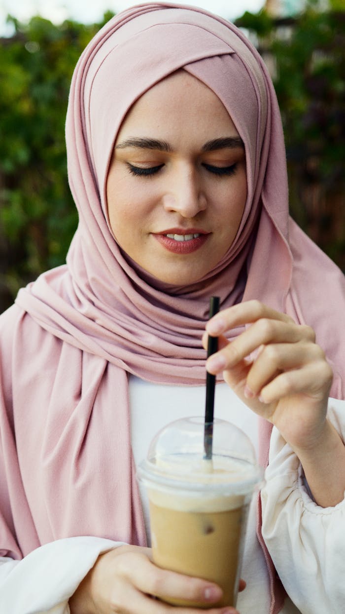 A young Muslim woman in a pink hijab enjoying a cold iced coffee drink outdoors.